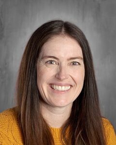 Smiling woman with long, dark hair wearing a yellow sweater, set against a gray background.