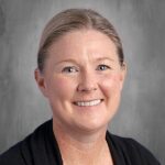 Smiling woman with light brown hair, wearing a black cardigan, against a gray background.