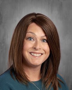 Smiling person with long brown hair, wearing a teal sweater, against a gray background.