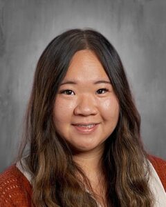 A smiling woman with long, wavy hair, wearing a brown sweater, poses against a neutral background.