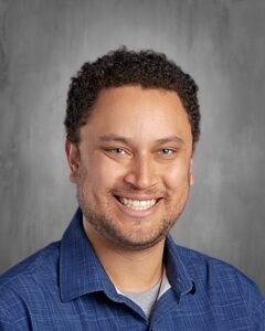 Hombre sonriente con cabello rizado y camisa azul, sobre un fondo gris.