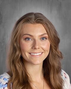 A smiling woman with long, wavy brown hair and blue eyes, wearing a floral top, against a gray background.