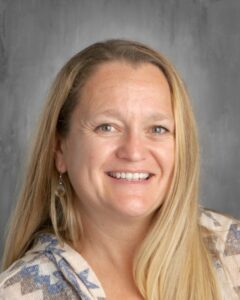 Smiling woman with long blonde hair, wearing a patterned sweater, against a gray background.