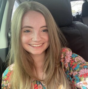 A smiling young woman with long, straight hair wearing a colorful floral top, seated in a car.
