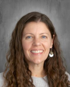 Smiling woman with long, curly brown hair, wearing a light sweater and earrings, against a gray background.