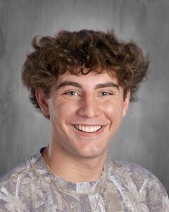 Young man with curly hair smiles brightly, wearing a camouflaged shirt against a gray background.