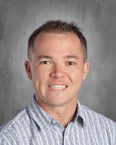 A smiling man with short hair wearing a striped button-up shirt, against a gray background.