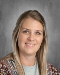 Smiling woman with long, light brown hair, wearing a patterned cardigan, against a gray background.