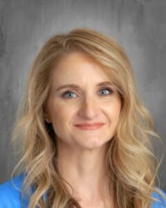 A smiling woman with long, wavy blonde hair and blue eyes, wearing a blue shirt, against a gray background.
