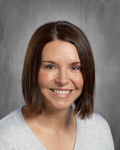 Smiling woman with shoulder-length brown hair, wearing a light gray sweater, against a gray background.