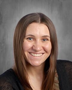Smiling woman with long brown hair and blue eyes, wearing a black cardigan, against a gray background.