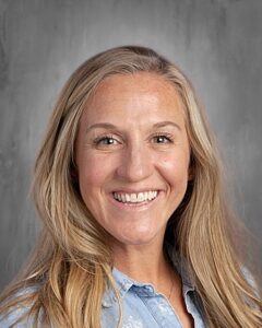 A smiling woman with long, wavy blonde hair, wearing a light blue shirt, against a gray background.