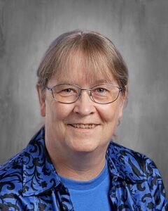 Smiling individual with glasses and long hair, wearing a blue shirt and patterned jacket, against a gray background.