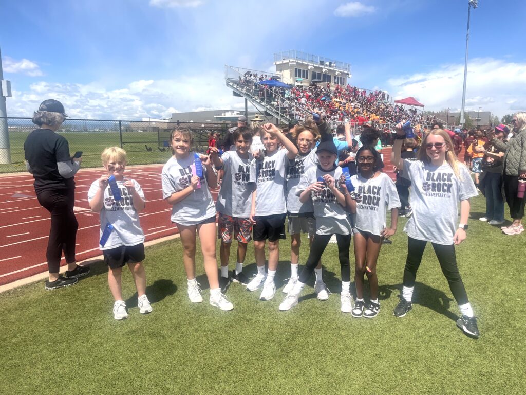 A group of seven children in gray t-shirts are posing with medals at a track event, with spectators in the background.