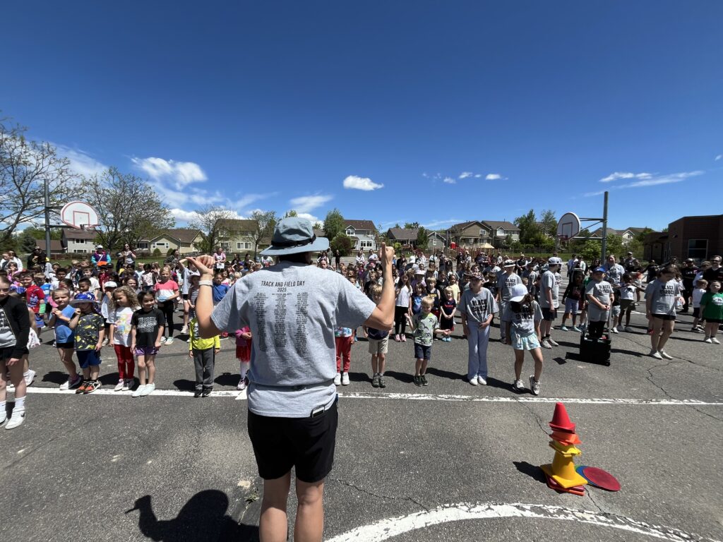 A crowd of children gathers on a playground for Track and Field Day, with a leader addressing them under a clear blue sky.