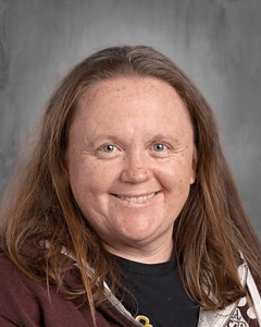 Smiling individual with long brown hair, wearing a dark hoodie, against a gray background.