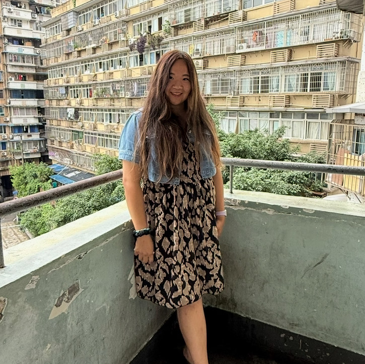 A person with long hair stands on a balcony, smiling, with a cityscape of apartment buildings in the background.