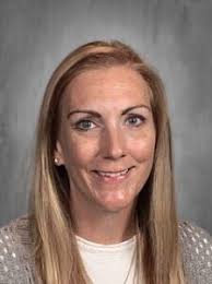 Headshot of a smiling woman with long blonde hair, wearing a light sweater and necklace, set against a gray background.