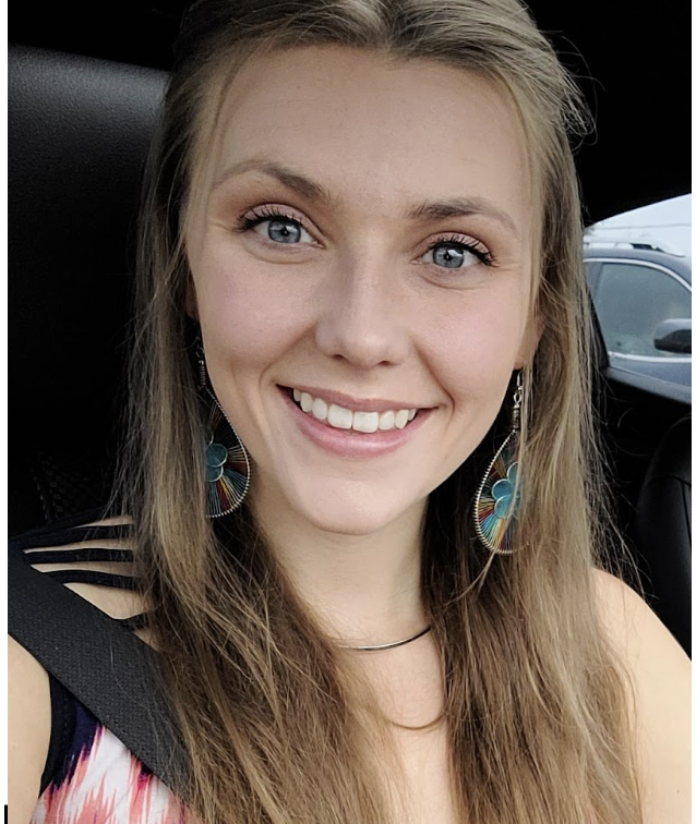 Smiling young woman with long hair, wearing colorful earrings, seated in a car with a seatbelt on.
