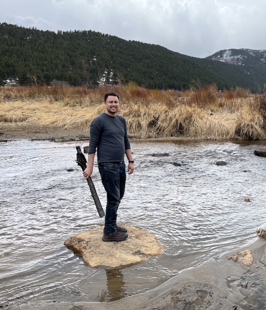 A person stands on a rock in a river, holding a stick, with grassy banks and mountains in the background.