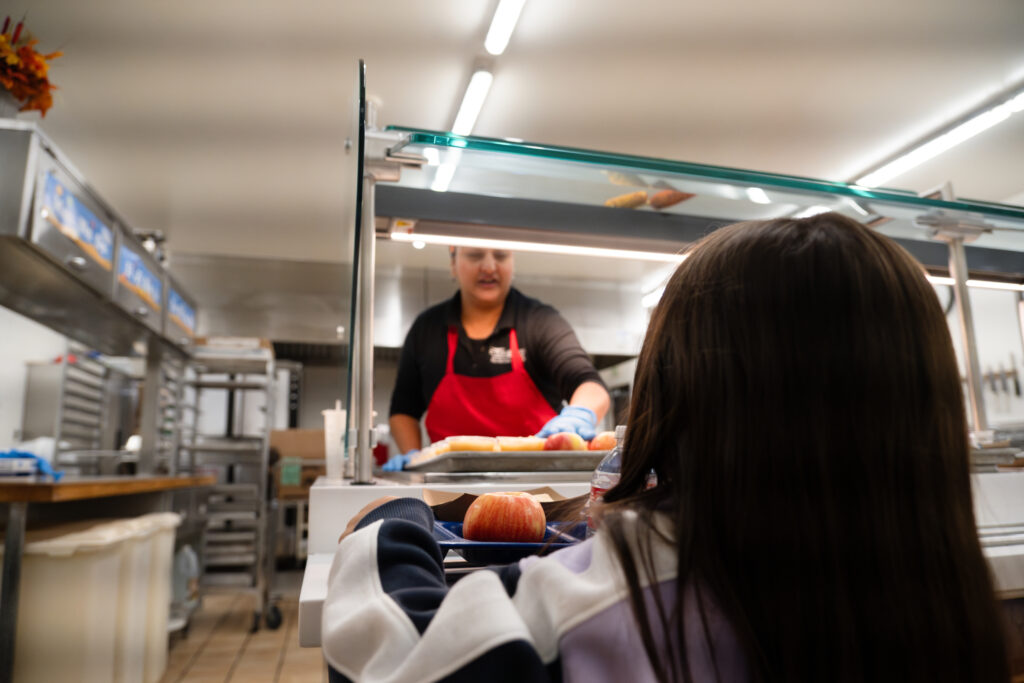 Un niño espera en el mostrador de una cafetería mientras un empleado sirve comida, incluida una manzana en una bandeja.