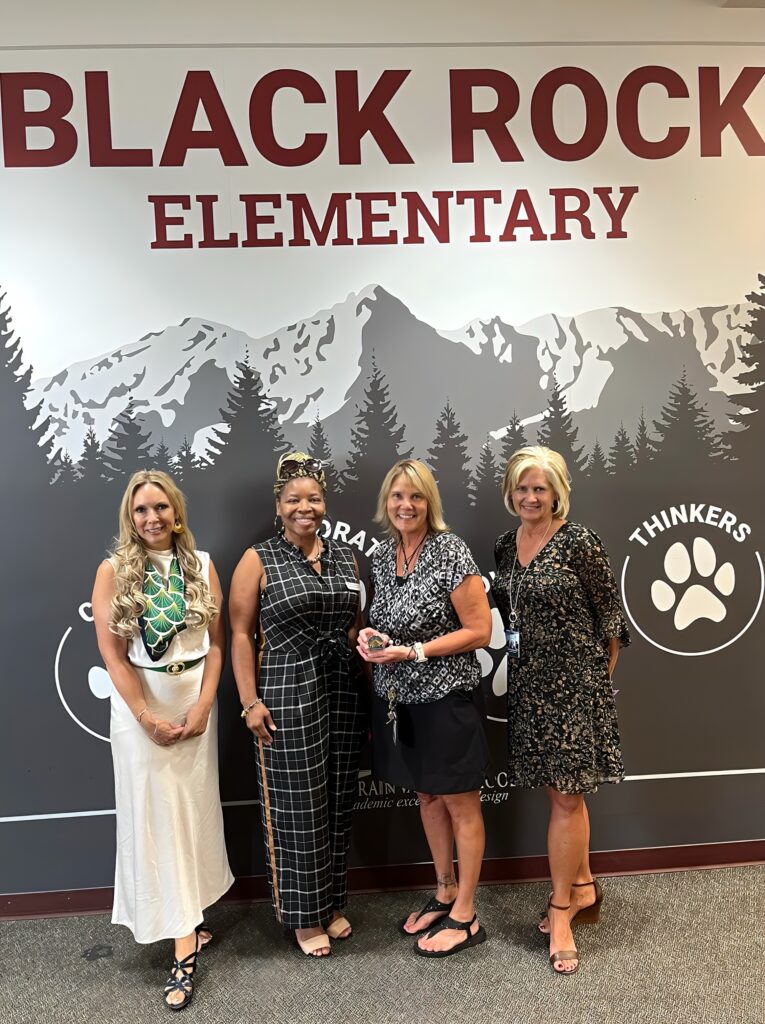 Cuatro mujeres sonríen y posan juntas ante un mural de temática montañesa en la escuela primaria Black Rock .