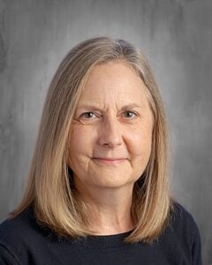 A woman with shoulder-length brown hair smiles at the camera, wearing a dark sweater against a neutral background.