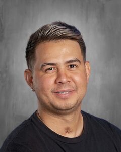 Smiling man with short hair and a light beard, wearing a black shirt, against a gray background.