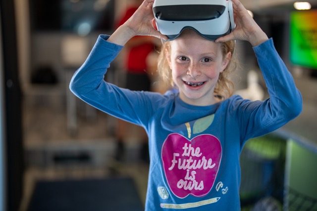 A young girl with red hair smiles while holding a virtual reality headset, wearing a blue shirt with an apple graphic.