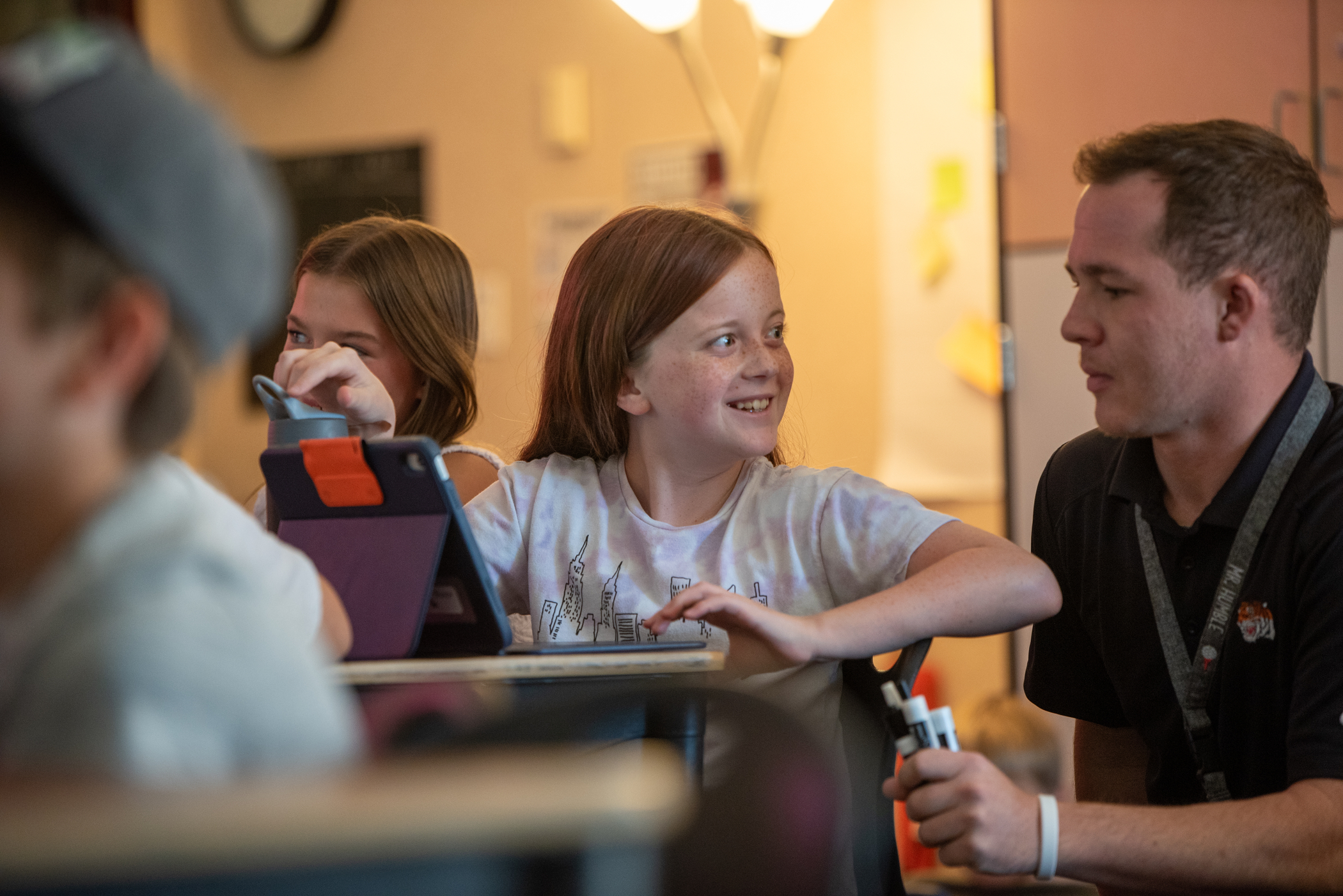 A girl at a classroom desk smiles at a teacher who is crouched beside her, holding dry erase markers.